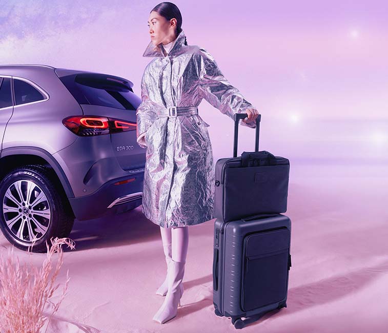 Eine Person trägt einen silbernen, glänzenden Mantel mit Gürtel und hält einen schwarzen Koffer mit Teleskopgriff. Im Hintergrund steht ein Auto.